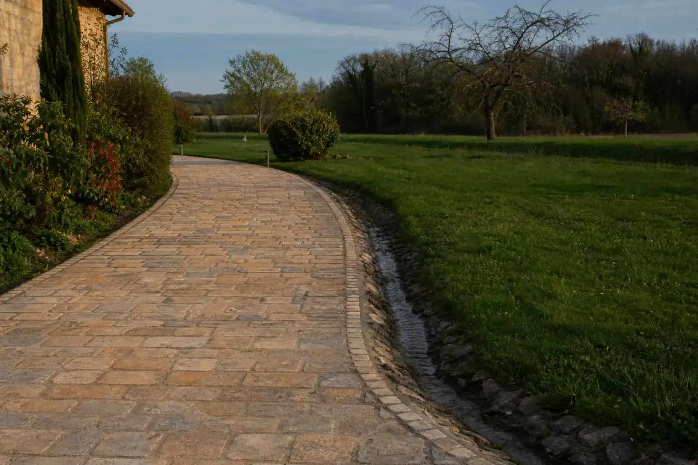 Allée de pavés en teintes dorées bordée d'une bordure pierre le long d'une pelouse, terrain frais du bocage bressan, drain visible en rive, ciel semi-continental.