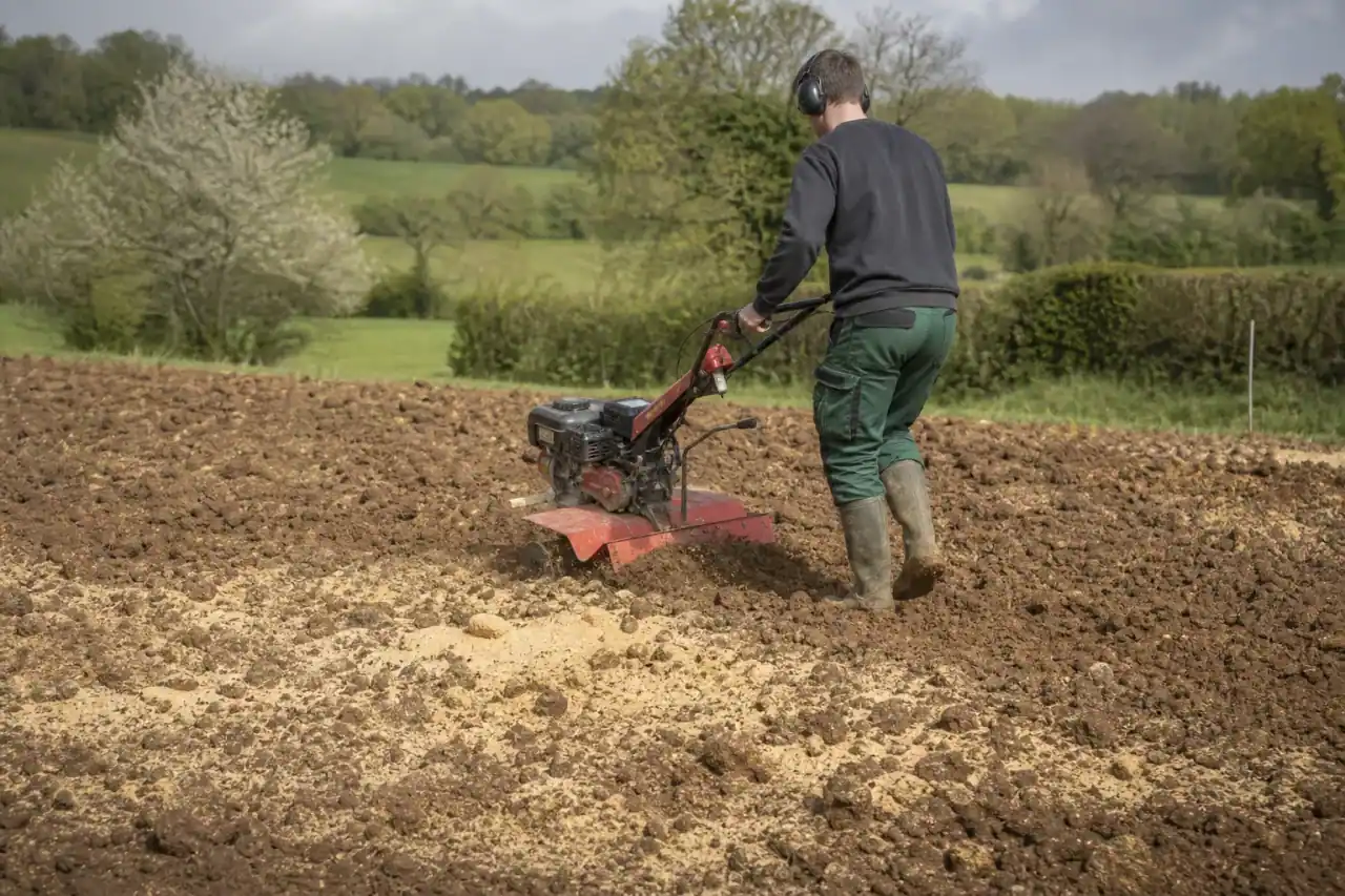 Paysagiste passant le rotavator pour décompacter une terre lourde et fraîche avant semis, mottes d'argile retournées, apport de sable visible au sol, terrain de bocage humide en fond.