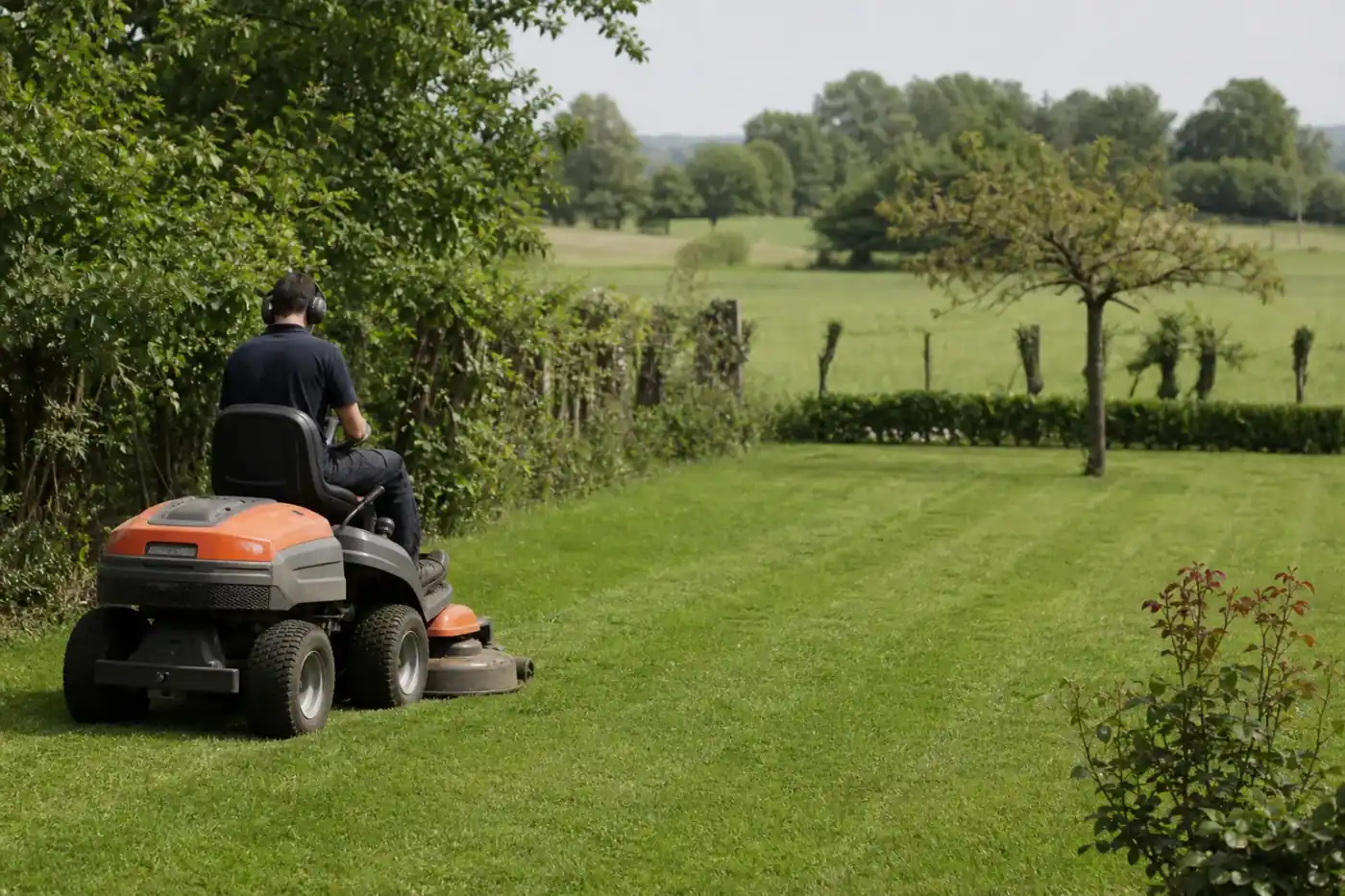 Jardinier tondant une grande pelouse de plaine avec une tondeuse autoportée, terrain plat et haies bocagères de la campagne bressane en arrière-plan.