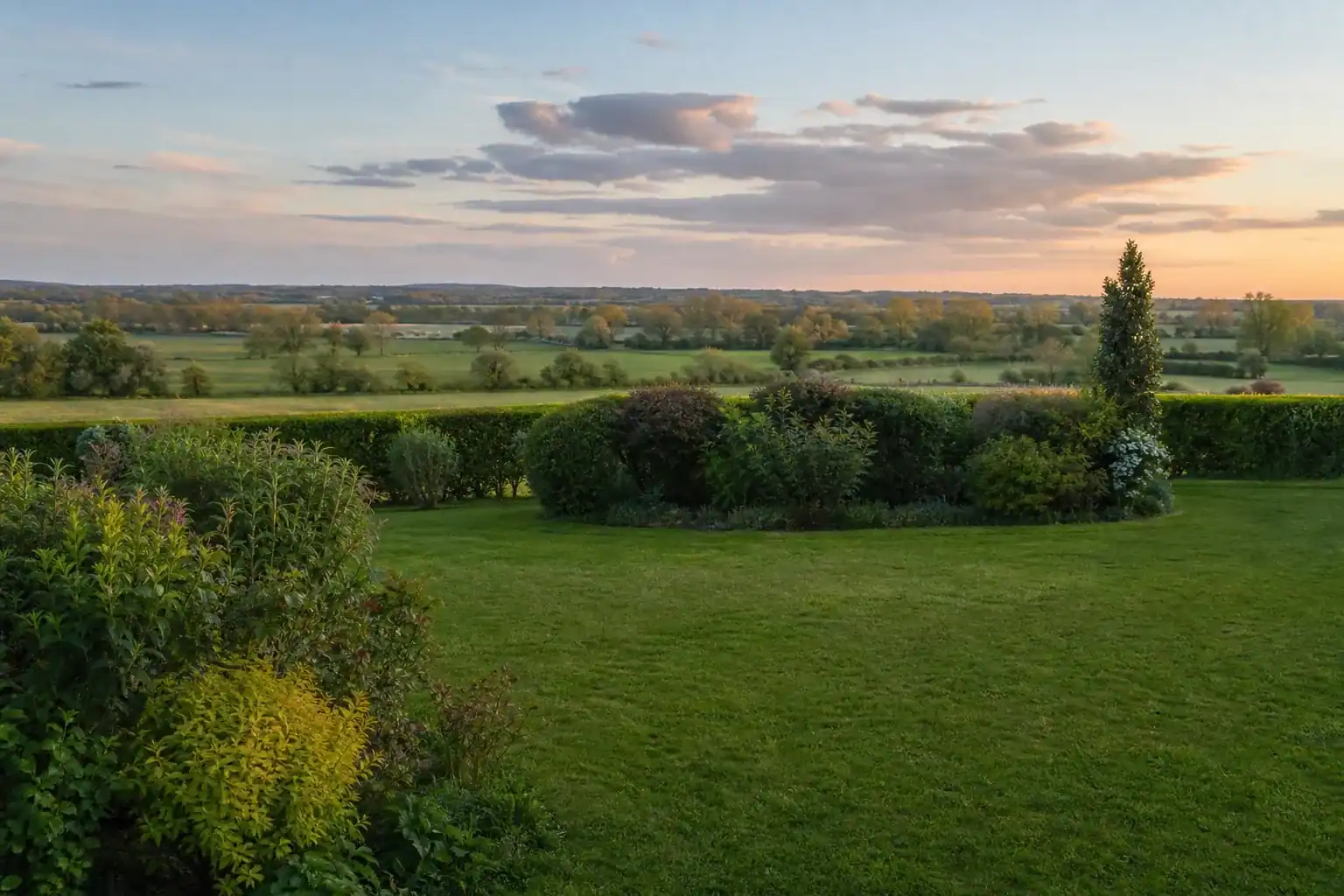 Jardin de pavillon en pleine campagne bressane à Attignat : grande pelouse verte et drue bordée d'une haie bocagère taillée, massifs d'arbustes qui structurent l'espace, campagne de prairies et de haies visible en arrière-plan, ciel de fin de journée aux nuages doux, cadrage large légèrement en plongée montrant l'ampleur du terrain rural.