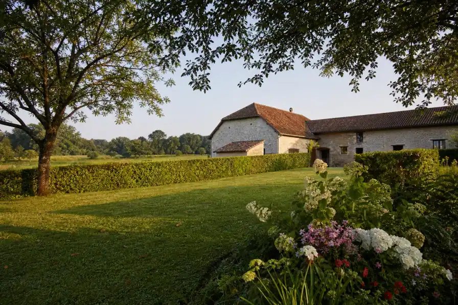 Jardin de campagne bressane sur un grand terrain à Viriat: une haie bocagère taillée abrite une pelouse dense et un massif généreux, une ferme bressane à toit de tuiles en arrière-plan, lumière douce de fin d'après-midi, prairies et haies à l'horizon, cadrage large qui laisse respirer l'espace.