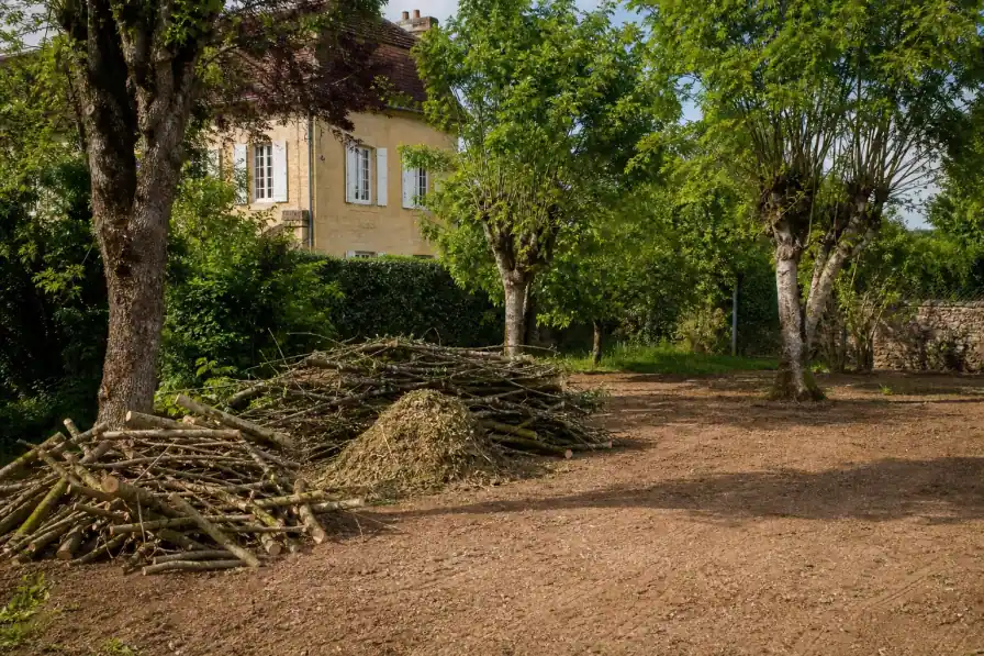 Branches taillées rassemblées et broyat prêt à évacuer, jardin dégagé et sol ratissé après le passage, maison en arrière-plan.