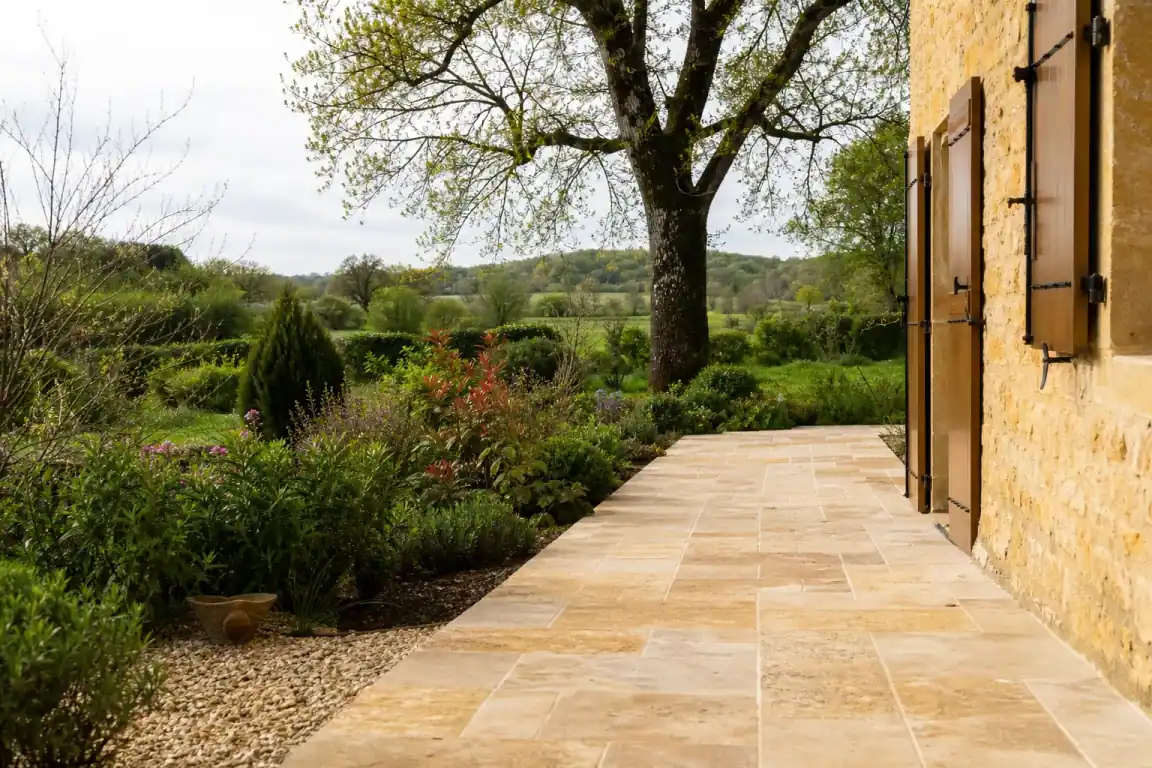 Détail d'une terrasse en pierre chaude le long d'une façade en pierre dorée, joints réguliers, pente légère visible, jardin et haie bocagère autour.
