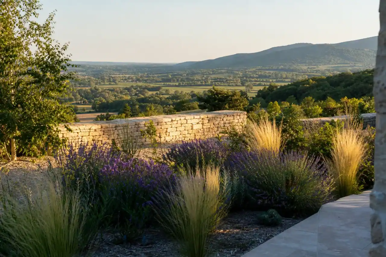 Photo d'un jardin en léger dévers à Ceyzériat au pied du Revermont, massif de lavandes et de graminées sur un sol caillouteux bien drainé, muret de pierre calcaire clair, vue ouverte sur la plaine bressane en contrebas, coteaux boisés du Jura à l'arrière-plan, lumière chaude de fin d'après-midi, cadrage large depuis la terrasse.