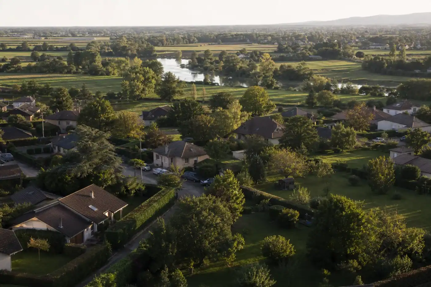 Vue aérienne basse d'un quartier pavillonnaire de la couronne de Bourg-en-Bresse : jardins verts contigus, haies bocagères taillées, la Reyssouze et les prairies de la Bresse en arrière-plan, lumière de fin d'après-midi.