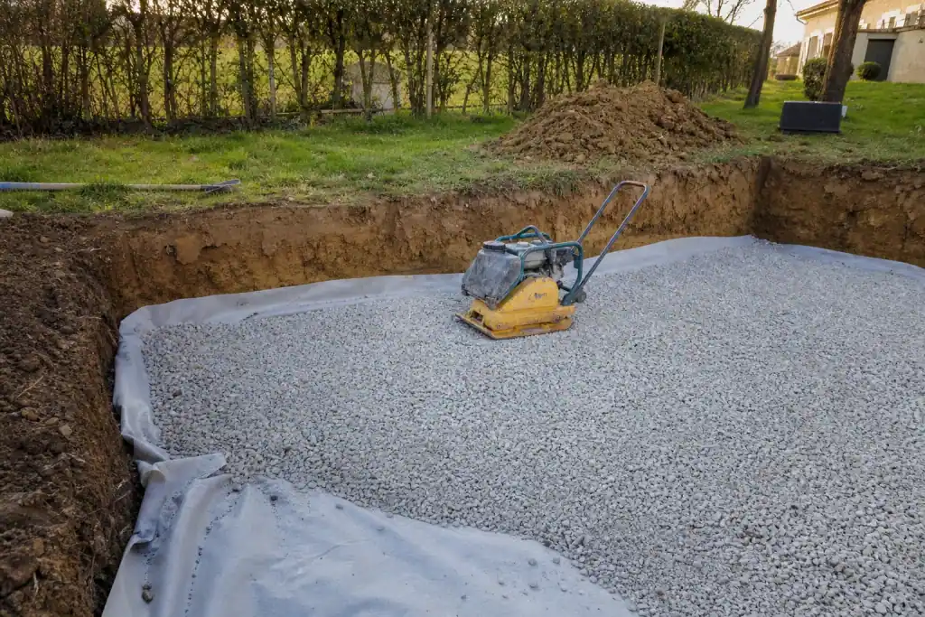 Gros plan sur une fondation de terrasse en cours : décaissement dans une argile brune de Bresse, géotextile et grave concassée compactée, plaque vibrante posée, chantier propre par temps clair.